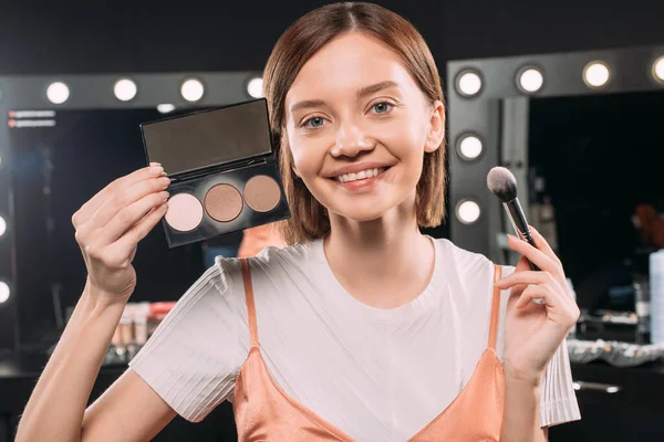 Hermosa mujer sonriente sosteniendo cepillo cosmético y maquillaje conjunto en el estudio de fotos - foto de stock