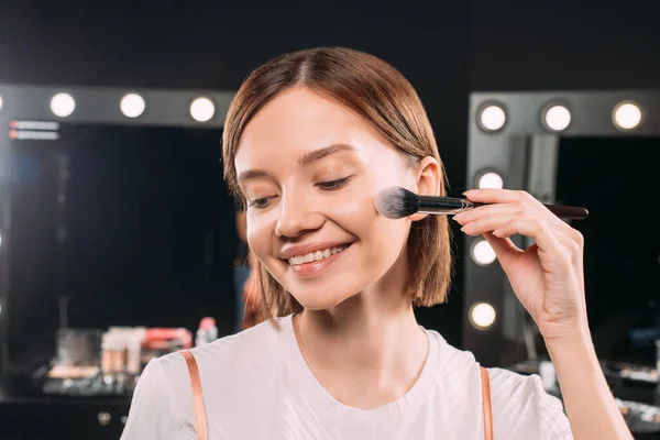 Bella donna sorridente che applica polvere per il viso con pennello cosmetico in studio fotografico — Foto stock