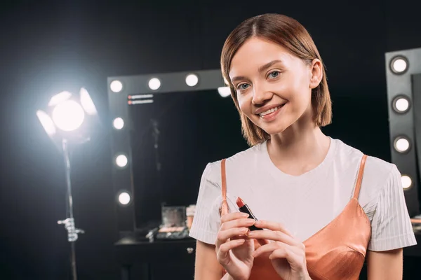 Hermosa mujer sonriente sosteniendo lápiz labial rojo en estudio de fotos — Stock Photo