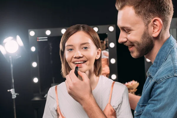 Artista de maquillaje guapo aplicando lápiz labial rojo en hermosa modelo sonriente - foto de stock