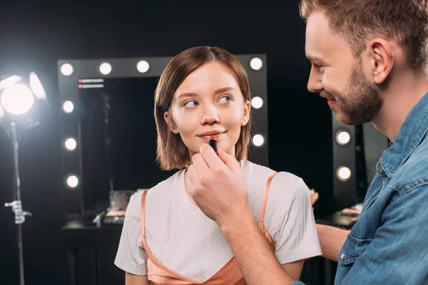 Sonriente artista de maquillaje aplicando lápiz labial rojo en atractiva modelo joven en el estudio de fotos - foto de stock