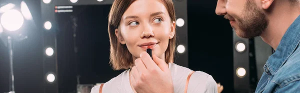 Imagen panorámica de una hermosa modelo mirando a un artista de maquillaje sonriente sosteniendo lápiz labial rojo en un estudio fotográfico - foto de stock
