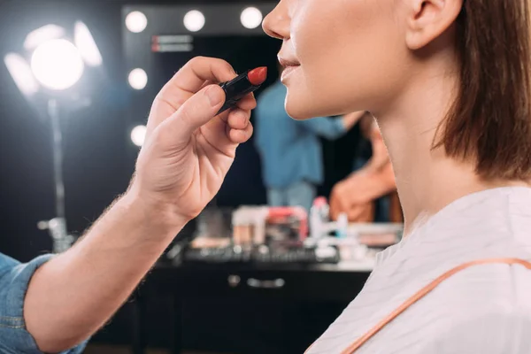 Vista ritagliata del truccatore che tiene il rossetto rosso vicino al giovane modello in studio fotografico — Foto stock