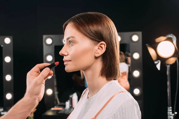 Vista lateral del artista de maquillaje sosteniendo lápiz labial rojo mientras hace el maquillaje a hermosa modelo en el estudio de fotos - foto de stock