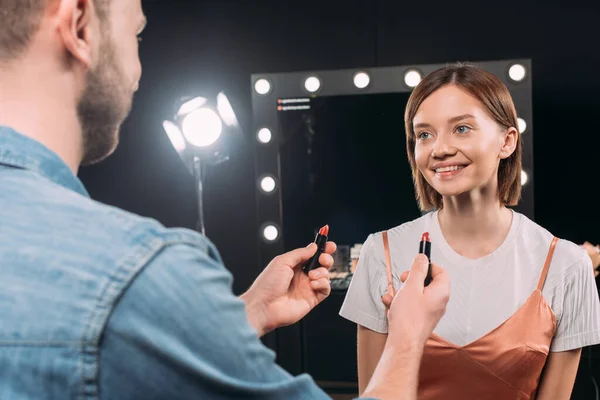 Focus selettivo dell'artista del trucco che tiene i rossetti vicino al modello sorridente in studio fotografico — Foto stock