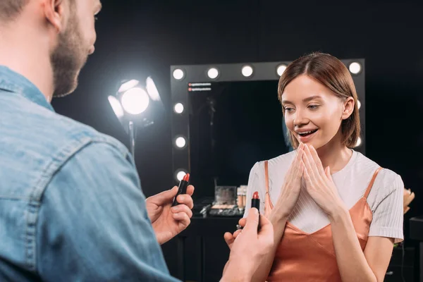 Enfoque selectivo del maquillador sosteniendo lápices labiales modelo casi sorprendido en estudio de fotos - foto de stock