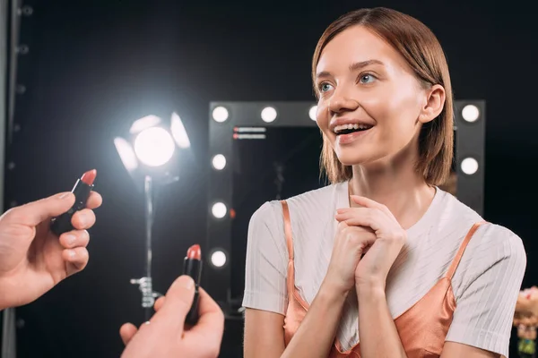 Maquillaje artista sosteniendo lápiz labial cerca de modelo positivo en estudio de fotos - foto de stock