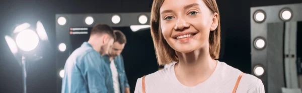 Panoramaausschnitt einer schönen lächelnden Frau, die in die Kamera blickt, während Make-up-Artist neben Spiegel im Fotostudio steht — Stockfoto