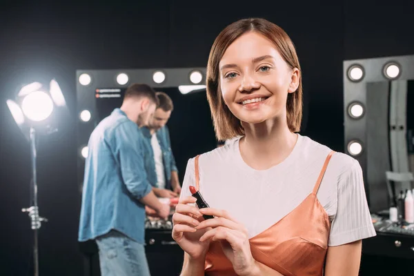 Bella modella sorridente che guarda la fotocamera mentre tiene il rossetto rosso in studio fotografico — Foto stock