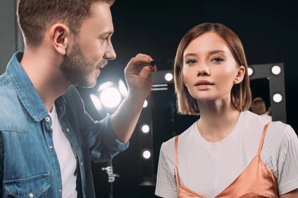 Hermoso artista de maquillaje sosteniendo rímel cerca de modelo atractivo mirando a la cámara en el estudio de fotos - foto de stock