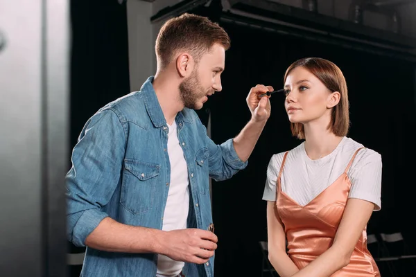 Artista de maquillaje guapo aplicando rímel en hermosa modelo joven en el estudio de fotos — Stock Photo