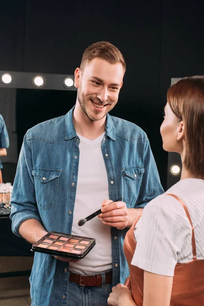 Selective focus of handsome makeup artist smiling at model while holding cosmetic brush and set of blush — Stock Photo