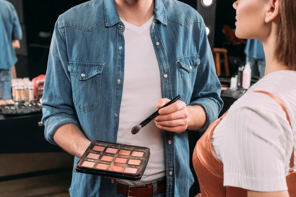 Vista ritagliata di trucco artista tenendo insieme di rossore e pennello cosmetico vicino al modello in studio fotografico — Foto stock