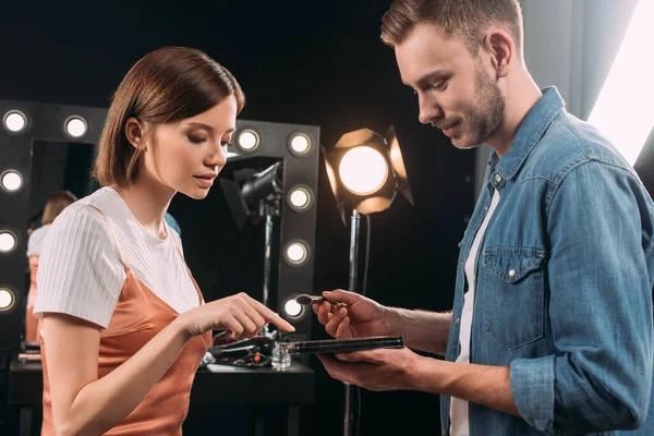 Vista lateral de hermosa modelo apuntando en el conjunto de maquillaje cerca de artista de maquillaje guapo con cepillo cosmético - foto de stock