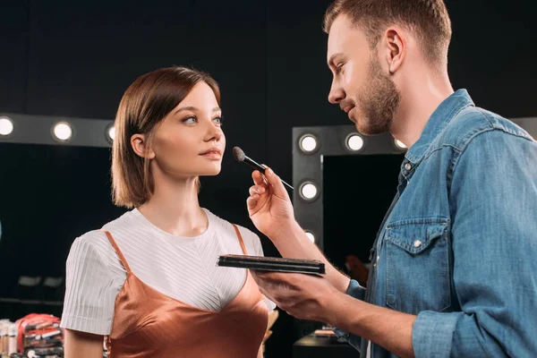 Artista de maquillaje guapo sosteniendo conjunto de maquillaje y cepillo cosmético cerca de la hermosa mujer joven en el estudio de fotos - foto de stock