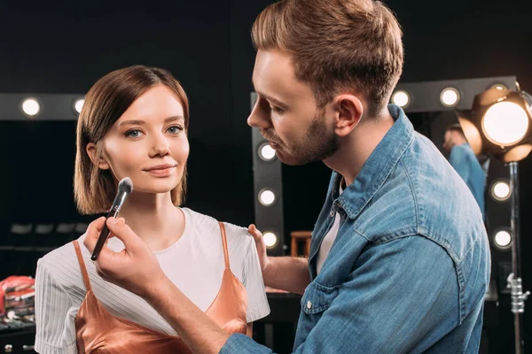 Artista de maquillaje guapo aplicando polvo facial en modelo atractivo en estudio fotográfico — Stock Photo