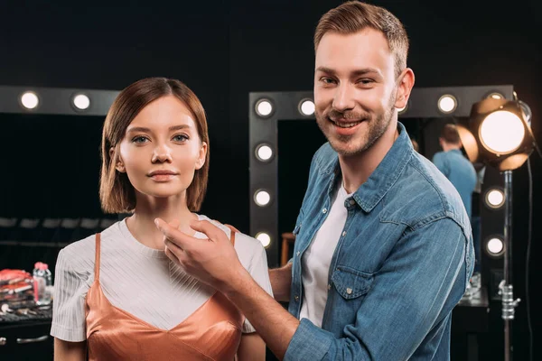 Handsome makeup artist smiling at camera near beautiful model in photo studio — Stock Photo