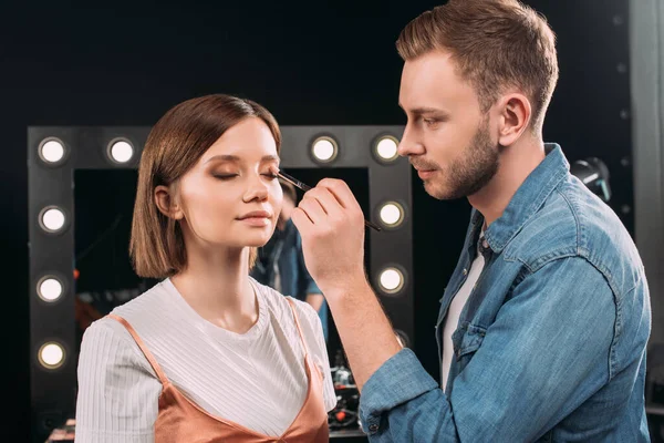 Artista de maquillaje guapo aplicando sombra de ojos en modelo en estudio fotográfico — Stock Photo