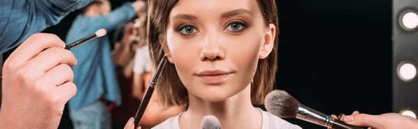 Panoramic orientation of makeup artists holding cosmetic brushes near beautiful model in photo studio — Stock Photo