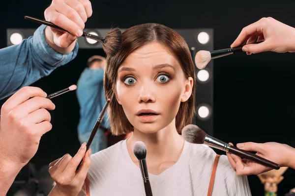 Makeup artist holding cosmetic brushes near shocked model looking at camera in photo studio — Stock Photo