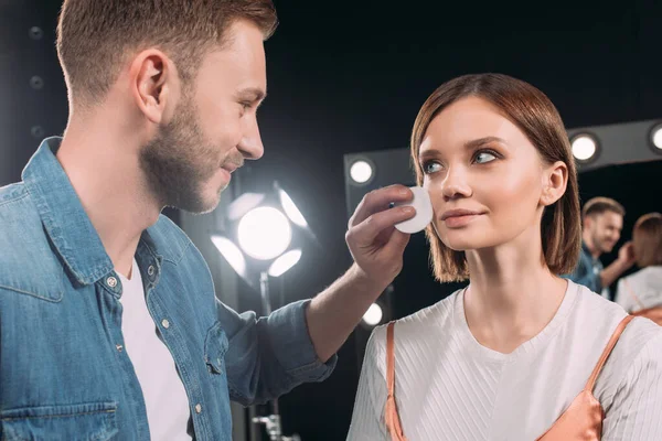 Sonriente artista de maquillaje sosteniendo almohadilla de algodón cerca de la mejilla de hermosa modelo en el estudio de fotos - foto de stock