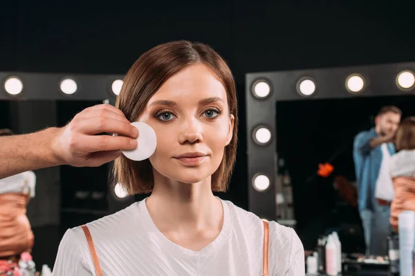 Maquiagem artista segurando almofada de algodão perto da bochecha do modelo atraente no estúdio de fotos — Fotografia de Stock