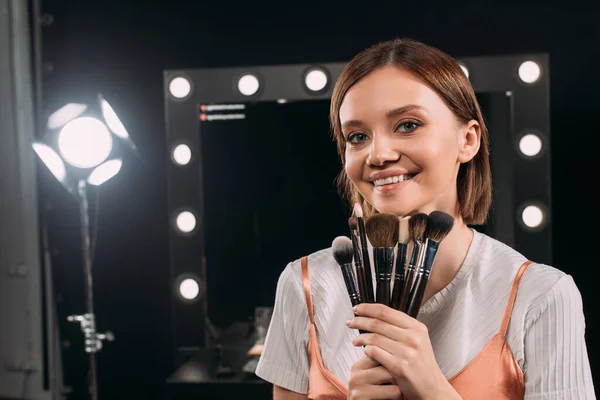 Hermosa modelo sonriendo a la cámara mientras sostiene cepillos cosméticos en el estudio de fotos - foto de stock