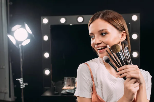 Schönes junges Model lächelt, während sie Kosmetikpinsel im Fotostudio hält — Stockfoto