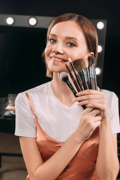 Mulher atraente segurando escovas cosméticas e sorrindo para a câmera no estúdio de fotos — Fotografia de Stock