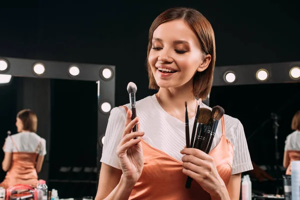 Positive model holding cosmetic brushes near mirrors at background in photo studio — Stock Photo