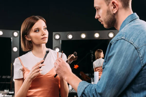 Maquillaje artista sosteniendo cepillos cosméticos y mirando hermosa modelo en el estudio de fotos - foto de stock