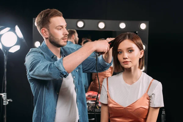 Artista de maquillaje guapo usando rizador en el cabello de hermosa modelo en estudio de fotos - foto de stock