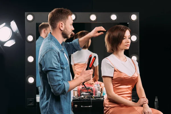 Maquillaje artista sosteniendo rizador y tocando el pelo de modelo en estudio de fotos - foto de stock