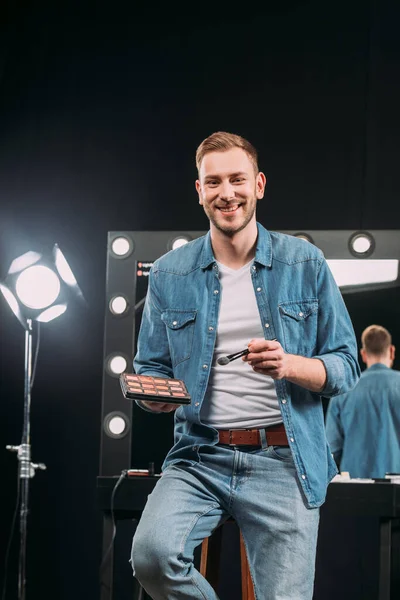 Hermoso artista de maquillaje sosteniendo conjunto rubor y cepillo cosmético mientras sonríe a la cámara en el estudio de fotos - foto de stock