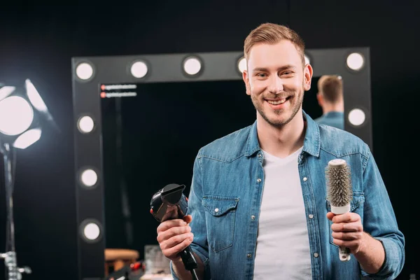 Guapo artista de maquillaje sosteniendo cepillo de pelo y secador de pelo y sonriendo a la cámara en el estudio de fotos - foto de stock