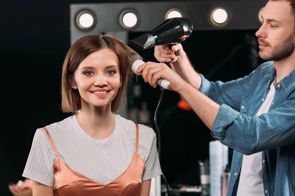 Handsome makeup artist doing hairstyle with hairbrush and hair dryer on smiling model in photo studio — Stock Photo
