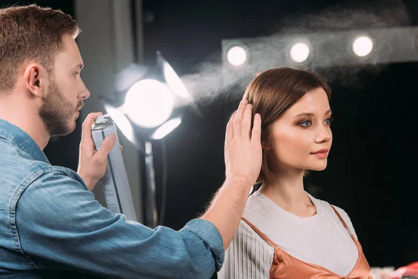 Beau maquilleur utilisant la laque à cheveux tout en faisant coiffure à beau modèle en studio photo — Photo de stock