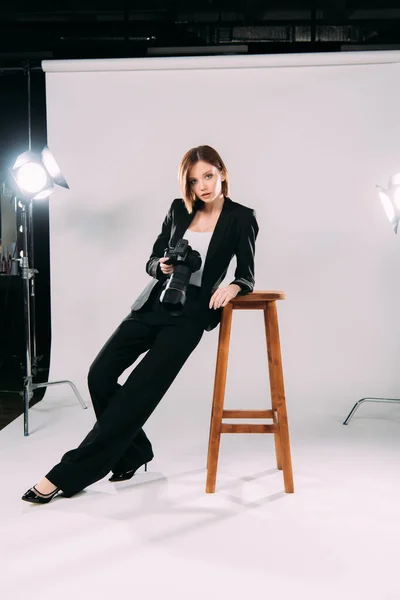 Beautiful model in formal wear holding digital camera near chair in photo studio — Stock Photo