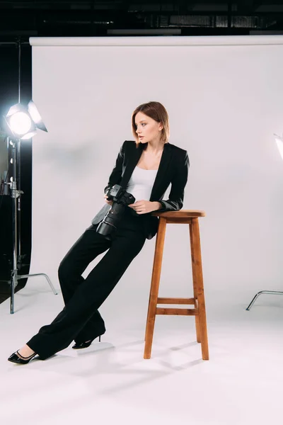 Elegante modelo con cámara digital cerca de la silla en el estudio de fotografía - foto de stock