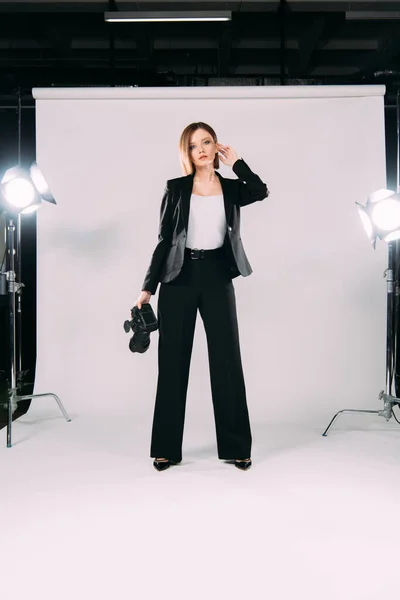 Beautiful model in formal wear holding digital camera near floodlights in photo studio — Stock Photo