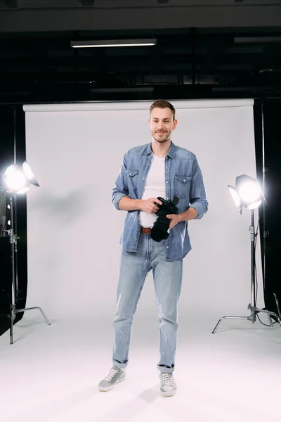 Hermoso fotógrafo sosteniendo la cámara digital y sonriendo a la cámara en el estudio de fotografía - foto de stock