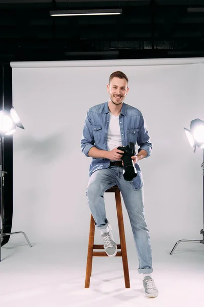 Красивый фотограф улыбается в камеру, держа цифровую камеру на стуле в фотостудии — стоковое фото