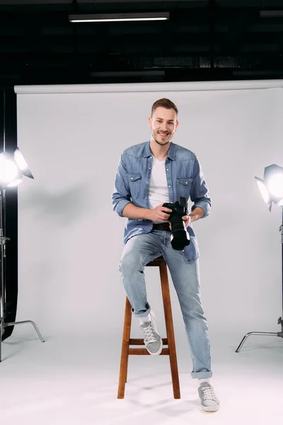 Beau photographe souriant à la caméra tout en tenant l'appareil photo numérique sur la chaise dans le studio de photo — Photo de stock