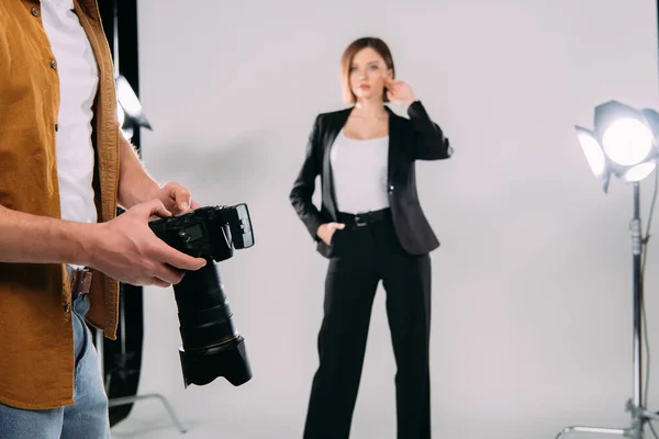 Mise au point sélective du photographe tenant un appareil photo numérique tout en modèle élégant posant en studio photo — Photo de stock
