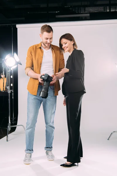 Bella modella sorridente che punta con mano a esposizione di macchina fotografica digitale vicino a fotografo bello in studio di fotografia — Foto stock