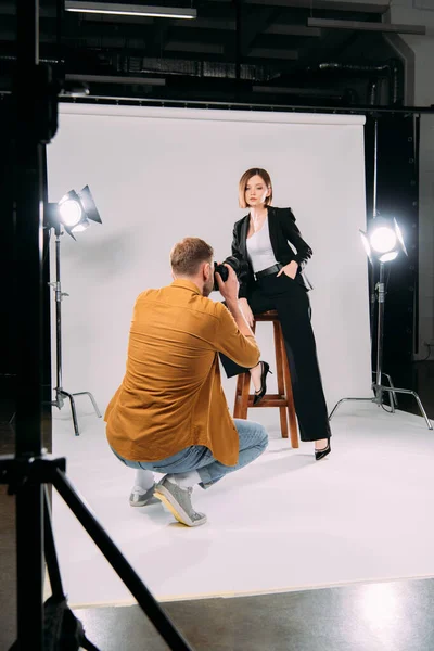 El foco selectivo del fotógrafo que trabaja con el modelo de estilo en la silla en el estudio de fotos - foto de stock