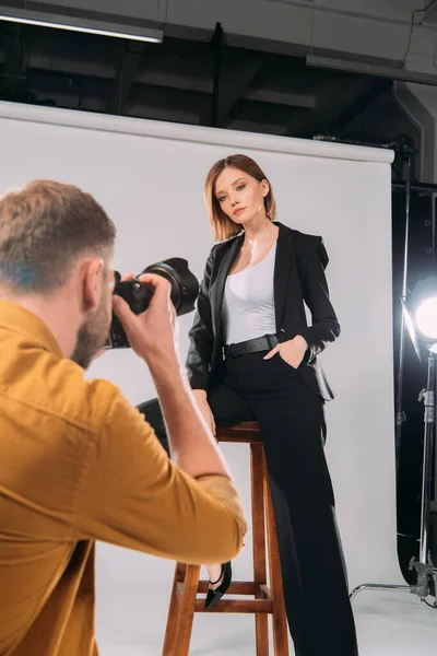 Selective focus of elegant model posing on chair at photographer in photo studio — Stock Photo