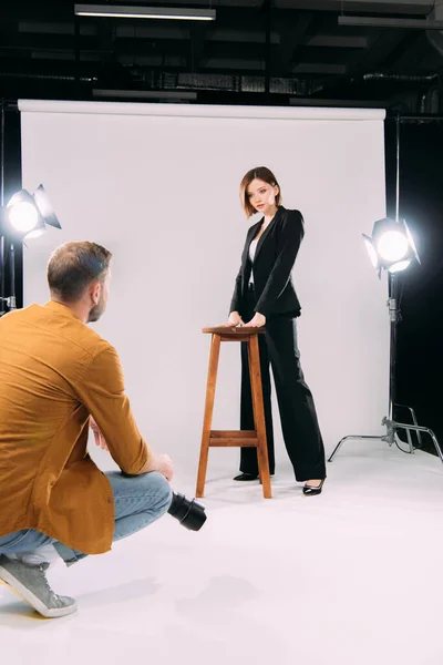 Beau modèle debout près de la chaise et regardant photographe avec appareil photo numérique en studio photo — Photo de stock