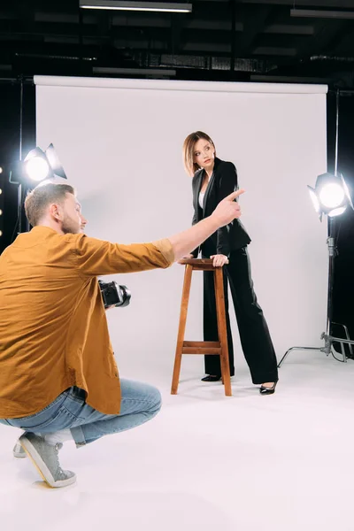 Fotograf zeigt mit Finger auf schönes Model in Stuhlnähe im Fotostudio — Stockfoto