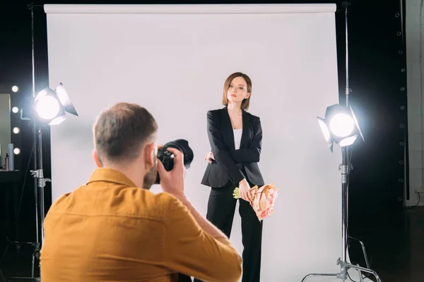 El foco selectivo del modelo de estilo con el ramo posando al fotógrafo con la cámara digital en el estudio fotográfico - foto de stock
